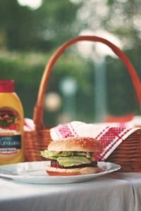 Picnic table set with food, drinks, and a colorful towel on a backyard deck with a bottle of sunscreen and a sandwich