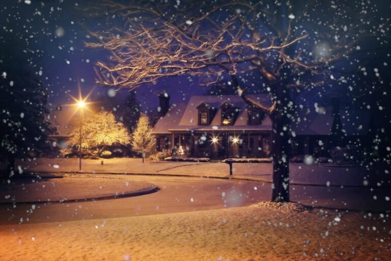 Snow falling on a residential street at night, with houses and trees illuminated by bright outdoor lighting